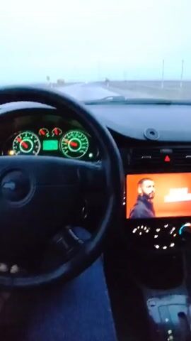The video shows a car interior view through the windshield while driving on a road. The driver is wearing jeans. The dashboard has a digital speedometer and a screen displaying a man's portrait. Music is playing. The sky is overcast. A truck passes in the distance. The camera pans to the left, showing a portion of the car's side mirror.