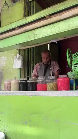 A man stands behind a brightly colored green counter in what appears to be a small food stall. He is wearing a dark apron over a light-colored shirt. In front of him, arranged in a line, are several large glass jars filled with various colorful ingredients. The jars contain items that look like beans, seeds, and possibly some kind of fruit or candy, in shades of pink, green, and black. The man is looking down at the counter, his hands obscured by the jars, but he appears to be engaged in preparing or serving something. To the right of the jars, a portion of a green ice shaver is visible. The stall has a rustic appearance with wooden beams overhead and a weathered wall behind the man. The overall impression is of a humble street food vendor preparing to serve customers.