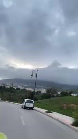 The video shows a cloudy day on a road overlooking a city. A white van is parked on the side of the road. A decorative street lamp is visible. The city is nestled in the foothills of a mountain, which is partially obscured by the clouds. The sun is trying to break through the clouds.