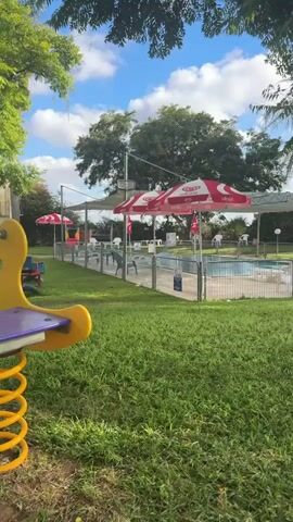 A panoramic video captures an outdoor scene. In the foreground, a lush green lawn fills the bottom of the frame. To the left, a portion of a yellow and purple playground structure is visible. The middle ground features a fenced-in swimming pool with blue water. Several red and white umbrellas are positioned around the pool area, providing shade. Stacked white plastic chairs are visible near a tree on the right side of the pool. A white plastic table and a yellow seesaw are also present on the lawn. The background consists of trees and a partly cloudy blue sky. The video is shot from a stationary perspective.