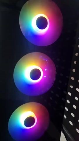 The video shows a close-up view of three spinning computer fans with rainbow-colored LED lighting. The fans are arranged vertically, one above the other, inside a dark enclosure. The lighting forms a ring around the center of each fan. The side of the enclosure is visible to the right, featuring a perforated pattern.