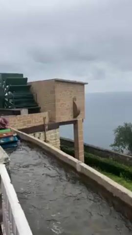 The video shows a scenic view of a coastal area with a water ride in the foreground. Initially, the camera is positioned near a water channel ride, focusing on a building with a water wheel. The scene then pans to the right, revealing the ocean and a distant coastline under a cloudy sky. As the camera continues to pan, a few trees are visible along the hillside, with a small boat seen in the distance. The final view showcases the coastline and the bay, revealing two small islands with a town nestled along the shoreline.