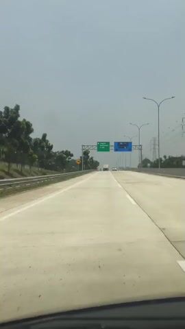 The video shows a point-of-view shot from a vehicle driving on a highway. The highway appears to be paved with concrete and has multiple lanes. On the left side of the road, there is a grassy median with trees and vegetation. On the right side, there are streetlights and high-voltage power lines. Overhead, a sign indicates that "Legok Karawaci" is 1 km away. There are other vehicles visible on the highway in the distance. The sky is overcast and a hazy gray. The camera appears to be mounted inside the vehicle, looking forward.