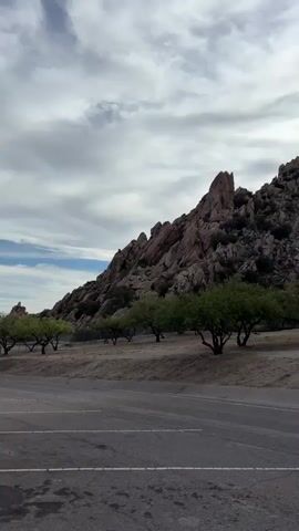 This is a vertical, panning video of a rocky hillside with trees in the foreground. The hillside is covered in large boulders and sparse vegetation. The trees are small and green, with rough bark. The ground in front of the trees is dry and sandy. A paved road is visible in the bottom portion of the video. The sky is overcast with some clouds. The sun is intermittently visible through the clouds. The video pans from left to right across the scene.