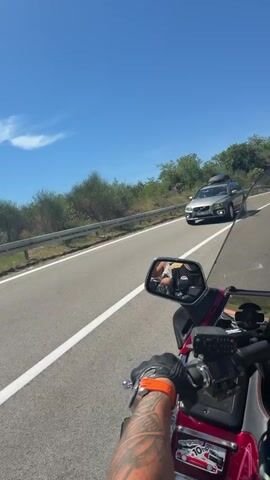 This is a first-person perspective video of someone riding a motorcycle. The rider's tattooed arm and gloved hand are visible on the handlebars. The motorcycle's windshield is in the foreground, and a side mirror reflects the rider and a passenger. The motorcycle is on a paved road with a white line marking the lane. A black car is passing the motorcycle in the opposite lane. To the left of the road is a rocky embankment covered with green bushes and trees. The sky is clear and blue.