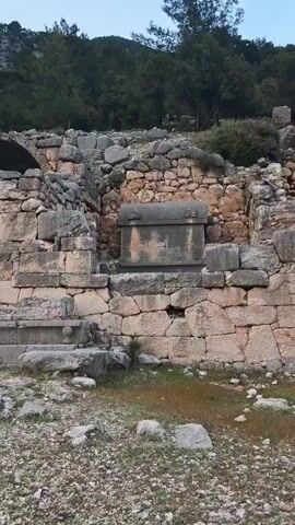 The video displays an ancient archaeological site characterized by substantial stone structures. A prominent, weathered, sarcophagus-like stone object, featuring a rounded lid, is centrally located on a raised platform, recessed into a larger stone wall. This wall is constructed from large, irregularly shaped blocks. Surrounding the sarcophagus are further ancient stone walls and scattered rubble, indicating extensive ruins.

In the foreground, the ground is uneven, covered with gravel, dirt, and patches of sparse grass. The background features a verdant hillside densely covered with pine-like trees, suggesting a Mediterranean landscape. The lighting appears soft and diffused, hinting at either early morning or late afternoon. No discernible text is visible on any of the structures. The scene evokes a sense of history and quiet solemnity within what are likely ancient ruins near Finike, Turkey.