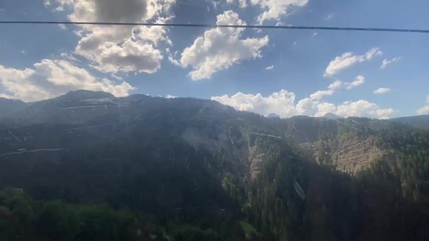 This media file captures a scenic view from a cable car ascending a mountain in Urtijëi - St. Ulrich in Gröden - Ortisei, Italy. The primary subject is the breathtaking mountain landscape, characterized by lush green forests covering the slopes and dramatic rocky outcrops. The sky is a vibrant blue, dotted with fluffy white clouds, suggesting a clear and pleasant day. Sunlight illuminates the scene, casting shadows and highlighting the textures of the mountainside.

A prominent feature is the cable car itself, with a section of the cabin visible in the foreground and along the bottom edge of the frame. The cable car appears to be in motion, moving upwards, as indicated by its position relative to the mountain. The activity taking place is the journey via cable car, offering panoramic views of the surrounding alpine environment.

Notable details include the time of day, which appears to be daytime, likely late morning or early afternoon, given the brightness of the sun. The weather is fair and sunny, conducive to outdoor activities and sightseeing. The location is clearly identified as the Dolomites region of Italy, known for its stunning mountain scenery. There are no people visible within the frame of the cable car cabin, suggesting the perspective is from inside or just outside an occupied car.

Text is visible on the side of the cable car cabin: "Val Gardena" and a logo which appears to be "G" with some stylized lettering underneath. This confirms the location as Val Gardena, a well-known valley in the Dolomites. The overall mood conveyed is one of awe and appreciation for the natural beauty of the mountains, with a sense of peaceful travel. The image is likely meant to evoke the experience of a scenic mountain excursion.