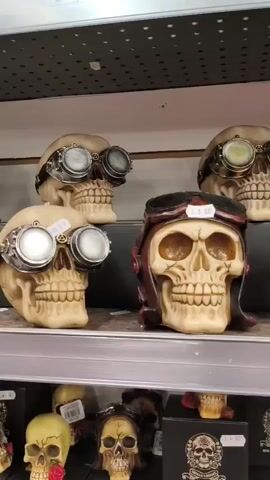 The video pans across a display shelf filled with decorative skull figurines. The skulls are depicted in various styles, some with steampunk-inspired goggles and helmets, others dressed as pirates with bandanas and hats. Some skulls are holding cigars or pipes. The figurines are positioned on shelves, and some are displayed in black boxes with skull emblems. The overall setting appears to be a retail environment, likely a novelty or gift shop. The lighting is even, suggesting an indoor setting with artificial lights. No people are visible in the video.
