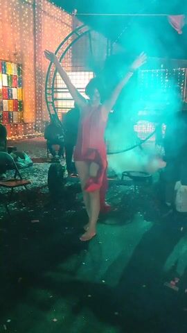 A barefoot woman in a short red dress with a train is posing with her arms up and later one hand on her head. There are several people standing and sitting in the background. There is a large metal wheel behind the woman. There is a lot of fog or smoke in the air, with a green and blue light shining on the scene. There are decorative lights in the background. The floor is littered with debris.