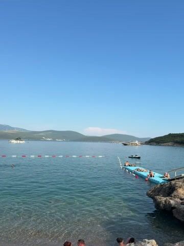 This is a vertical, eye-level, medium shot of a serene beach scene under a clear blue sky. The foreground features a pebbly shore where several people are partially visible, facing away from the camera. The water is a clear, turquoise hue, with gentle waves lapping at the shore. A red and white buoy line stretches across the water, separating the swimming area. In the middle ground, a bright blue floating dock extends from a rocky outcrop into the sea. Several individuals are lounging on the dock, sunbathing. A small boat is moored near the dock, and larger boats are visible in the distance. The background consists of a calm, expansive body of water and distant, tree-covered hills meeting the horizon under the vast expanse of the sky.