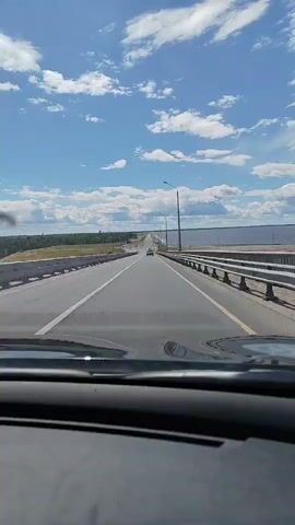 The video shows a point-of-view shot from inside a car driving on a highway. The highway is a multi-lane road with a guardrail on the right side. The road stretches out into the distance, with a few cars visible ahead. On the right side of the road, there is a large body of water, likely a lake or sea, and a flat, treeless landscape. On the left side of the road, there is more vegetation, appearing as fields and distant trees. The sky is blue with scattered white clouds, suggesting a pleasant, sunny day. The overall scene conveys a sense of travel and open road. The time of day appears to be daytime, likely afternoon, given the bright sunlight and clear sky. The location context provided is Ivanovo (Timiryazevskii S/S), Russia. No text is visible in the video.