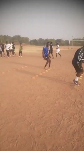 The video shows a group of young men doing agility drills on a dirt field. They are running and stepping between small orange cones arranged in a line. Some of the men are wearing soccer jerseys in different colors, including blue, black, and green, while others are in casual sportswear. A soccer goal is visible in the background. The field appears to be outdoors, with trees and vegetation in the distance under a bright sky.