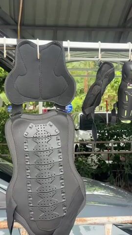 The video shows various pieces of protective gear for motorcycle riders hanging on a rack outdoors. These items include padded vests, knee pads, elbow pads, and helmets. The gear is predominantly black with some accents of neon green and red. A white and a brown helmet are visible. Some of the gear is hanging on metal hooks, while other pieces are clipped on. A grey jacket and a pink piece of fabric are also hanging on the rack. In the background, there are cars, trees, and a corrugated metal roof structure. The environment appears to be a covered outdoor area, possibly a garage or carport, with some greenery visible. There are no people present in the video.