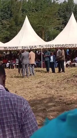 The video shows a gathering of people outdoors, likely for a formal event like a funeral or a community meeting. There are two large, white peaked tents set up with rows of people seated underneath. In front of the tents, a group of six people stand together, facing the crowd. The setting appears to be a grassy field with trees in the background. The weather seems fair and sunny. The camera angle is from behind another person, providing a partial view of the event.