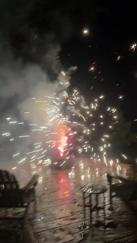 The video shows fireworks being set off on a paved patio at night. Two Adirondack chairs and a small stool are visible in the foreground. The fireworks are creating a bright light and smoke, with sparks flying outwards. The patio surface appears wet, possibly from rain. The scene is dark, with the exception of the light from the fireworks and some ambient light from the sky. There are no people visible in the video.