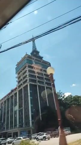 The video is filmed from inside a moving vehicle, likely a car, as evidenced by the reflections on the window. The camera is pointed upwards and to the right, capturing a tall, multi-story building with a distinctive tower at the top, resembling the Eiffel Tower. The building has a dark facade with white accents and multiple windows. There are power lines and poles visible in the foreground, suggesting an urban or suburban setting. The sky is blue and appears to be daytime. The vehicle passes by the building, and a pedestrian overpass is also visible. There are several cars parked on the street in front of the building. No people are clearly visible or identifiable in the video.