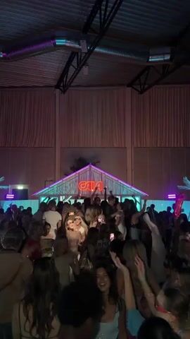 A large crowd of people, primarily young adults, dance enthusiastically in a dimly lit indoor venue under turquoise and purple stage lighting. The ceiling features exposed dark rafters and ventilation ducts. In the background, a small hut-like structure with a peaked roof and a glowing red neon sign spelling "GALA" serves as a focal point, flanked by two neon-outlined palm tree silhouettes. Green uplighting illuminates the wall behind the hut. Many attendees are smiling, laughing, raising their hands, and recording the event with smartphones. One person in the mid-left wears a white graduation cap with a blue ribbon. The overall atmosphere is lively and celebratory.