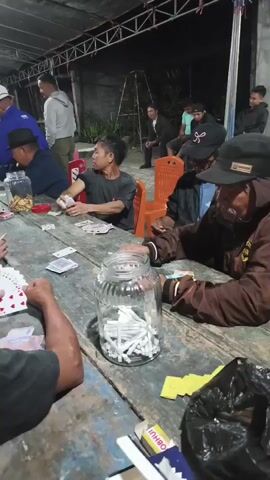 A group of men are gathered around a long wooden table playing cards. The scene appears to be outdoors under a covered structure, possibly at night, indicated by the artificial lighting. Most of the men are focused on their cards or the game, with some looking at each other. There are stacks of cards and money on the table, as well as several glass jars filled with cigarettes. One jar, prominently in the foreground, is filled to the brim with cigarettes. The atmosphere seems casual and social.