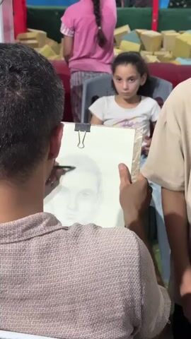 A man is drawing a portrait of a young girl. The artist is in the foreground, facing away from the camera, holding a clipboard with a drawing on it. He is using a pencil to draw the portrait. The girl is sitting in a chair in the middle ground, looking at the artist. She is wearing a white t-shirt. In the background, there are other people, including a girl in a pink shirt and a boy wearing a Spider-Man mask. The environment appears to be an indoor event, possibly a carnival or fair, with some soft padded structures visible in the background.