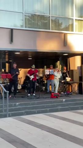 A live band is performing on a stage outdoors in front of a building. The band consists of five people: four men and one woman. Two men are playing guitars, one man is singing and playing guitar, one man is behind a drum set, and the woman is singing. The stage is set up on steps leading up to a building with large windows. There are various musical instruments and equipment on the stage, including guitars, keyboards, amplifiers, and microphones. The lighting suggests it is either late afternoon or early evening, with warm sunlight casting shadows. The ground in front of the stage is paved with geometric patterns.