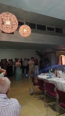 This is a wide shot of an indoor event, likely a party or reception. Two large, spherical light fixtures hang from the ceiling, emitting a warm glow. In the background, a group of people are gathered, and some are dancing. A woman in a sparkly blue-green dress and a man in a light-colored shirt and pants are in the center of the dance area, appearing to be dancing together. Other guests are clapping and watching them. To the right of the frame, a round table is set with white tablecloths, plates, bottles, and glasses, suggesting a dining area. A person wearing a striped shirt is visible in the foreground, facing away from the camera. The room has a light-colored floor and walls, with a ceiling that has a windowed band.