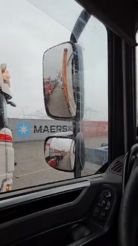 The video captures a first-person perspective from the driver's seat of a truck, looking out through the front windshield and the right-side mirrors. The scene outside is an industrial area, likely a port or container terminal, during what appears to be a rainy or overcast day, indicated by water droplets on the window and a generally dull sky.

Through the front windshield, a queue of various trucks, some blue, some white, and some red, is visible in the distance. Beyond them, large industrial cranes, typical of a shipping port, can be seen, partially obscured by the weather and the intervening vehicles. Several shipping containers, both red and blue, are also scattered throughout the area.

The right-side mirrors provide a clear reflection of the trucks directly behind and to the side of the viewer's vehicle, showing a continuous line of commercial trucks waiting. One large blue shipping container with the prominent white text "MAERSK" is clearly visible in the background through the mirror's reflection and also somewhat directly through the window.

Inside the cab, a black steering wheel with integrated controls is prominent in the foreground, along with part of the dashboard showing speedometer and tachometer gauges. The interior appears modern and well-maintained. The activity implied is waiting in a queue, possibly to load or unload cargo at the Maasvlakte Rotterdam port in the Netherlands, as suggested by the environment and location context.