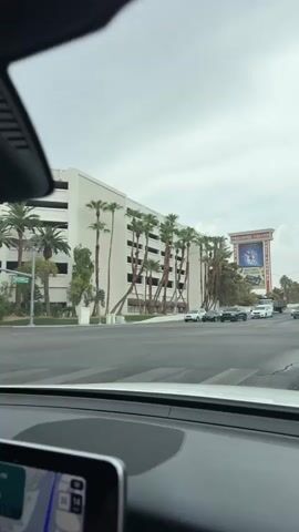 The video shows a street view from inside a car. The car is at an intersection, and traffic is moving slowly. In the background, there is a large building with palm trees in front of it. To the right of the building, there is a large billboard for "Treasure Island" which advertises "MYSTERE" and "FREE PARKING." The sky is overcast.