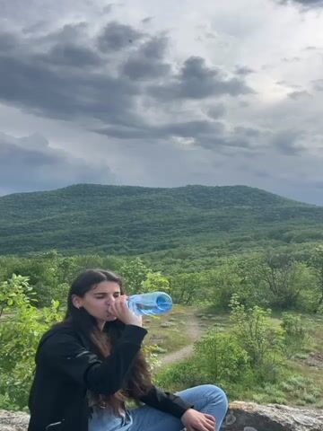 A young woman with long dark hair and wearing a black jacket and blue jeans is sitting on a rock outdoors. She takes a drink from a blue plastic water bottle. Behind her is a view of a green, forested hillside and a gray, cloudy sky. The woman looks at the camera after drinking.
