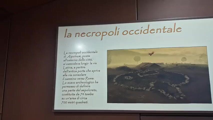 The video shows a close-up shot of a museum exhibit. The exhibit features a large rectangular display board with text and images. On the left side of the board, there is text in Italian, with the title "la necropoli occidentale" (the western necropolis) at the top. Below the title is a paragraph of text describing the necropolis. To the right of the text is a painting depicting an archaeological site with some circular stone structures and what appear to be ancient buildings or tombs. On the right side of the display board, there is a diagram showing different layers of soil and what appears to be a cross-section of an archaeological excavation. The diagram includes labels in Italian, such as "livelli di protezione" (protection levels), "suolo antico" (ancient soil), "terre rosse" (red earths), and "travertini" (travertine). Below these labels, there are representations of tombs, labeled "Tomba 6 del letto funerario" (Tomb 6 of the funerary bed) and "Tomba 7 della lastrona italica" (Tomb 7 of the Italic slab). Further to the right, there is a drawing of an "AUTOGRILL" sign and some trucks, suggesting a connection between the ancient site and modern infrastructure. The lighting in the video is somewhat dim, and there are some reflections on the display board. The background of the exhibit appears to be a dark, wooden panel. No people are visible in the video.