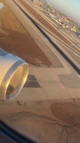 A video captures an aerial view of an airport runway as an airplane's engine is visible in the foreground, to the left. The runway is marked with white lines and numbers. In the distance, another airplane, predominantly pink and white, is maneuvering on a connecting taxiway or runway. The surrounding environment appears arid, with patches of brown earth and sparse vegetation. Buildings and infrastructure of the airport are visible in the background, along with other parked aircraft. The lighting suggests it is either sunrise or sunset, casting a warm glow on the scene. The perspective is from inside a passenger aircraft, looking out the window.