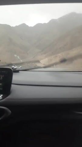 The view from inside a moving car shows a mountain landscape on a rainy day. The wipers are active.  The car's dashboard is visible with a touchscreen display. A decorative item, possibly a small bird, sits on the dashboard. Outside, the road is grey, and the mountains are muted in the distance. The sky is overcast.  There is a faint road marker visible on the right. The camera is stable, suggesting a smooth ride.