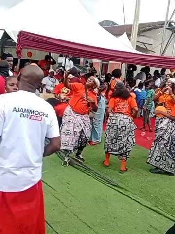 The video shows an outdoor event with a crowd of people. In the foreground, a man wearing a white t-shirt with the text "AJAMIMOGHA DAY 2026" and a red garment is facing away from the camera. Several women, mostly wearing orange tops and patterned skirts, are dancing and interacting in the middle ground. Some of them are holding orange balloons. A white tent with a maroon border is set up in the background. The ground appears to be covered in artificial turf.