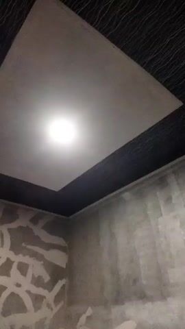 A video shows a dark room with a light fixture on the ceiling. The ceiling is white and rectangular, with a dark border around it. The walls are concrete and appear to be unfinished, with some abstract patterns painted on them.