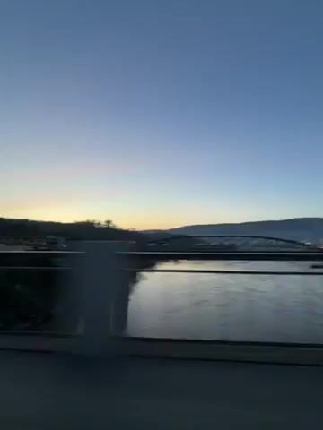 The video shows a bridge over water at dawn. In the distance is another bridge and some houses on a hill. Two cars pass on the bridge in the lower center of the frame. The sky is clear and ranges from dark to light blue.