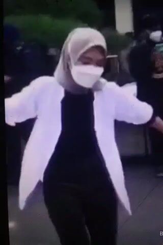 A young woman wearing a hijab, a white jacket, a black shirt, and dark pants is dancing. She is also wearing a white face mask. The woman is outdoors, and there are other people in the background. The video appears to be a short clip from a longer recording, with timestamps visible. The woman's movements are fluid and energetic as she dances.