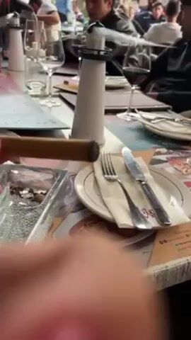 A person is lighting a cigar with a black torch lighter at a table in an outdoor restaurant. The table is set with silverware, wine glasses, a white decorative lighthouse, and an ashtray with what appears to be coffee beans in it. Other patrons are visible in the background, seated at nearby tables. The table surface has a colorful, patterned design.