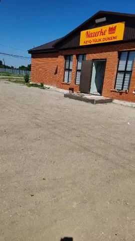 The video shows a brick building with a brown roof and a large yellow sign that reads "NAZERKE AZYO-TÜLİK DÜKENI". The building has several windows with black frames and a double door that is partially open. An air conditioning unit is mounted on the exterior wall. The ground in front of the building is covered in gravel and dirt. The sky is clear and blue. There are no people visible in the video, but there is a small black object that may be an animal near the entrance. The overall impression is of a small business or shop in a rural or semi-rural setting.