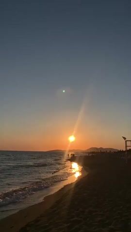 The video shows a sunset over a beach. The sun is a bright orange orb low on the horizon, casting a golden reflection on the water. The ocean waves gently lap the sandy shore. Silhouettes of people can be seen walking along the beach in the distance. The sky is a gradient of orange, pink, and blue, with a lens flare visible near the sun. The overall scene is serene and picturesque.