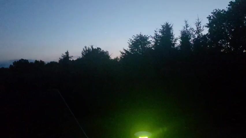 The video begins with a dark outdoor scene, possibly at dusk or dawn, with trees silhouetted against a twilight sky. A green light emanates from the bottom right corner of the frame. The scene then transitions to a view of a residential area at night. Several houses are visible, some with lights on. Streetlights illuminate the road and sidewalks. The sky is dark blue with some clouds. The video pans slightly, showing more of the houses and trees. The overall impression is of a quiet, nighttime suburban environment.