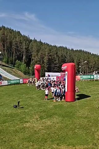The video shows a group of people gathered at the starting line of an outdoor event. There are two large red inflatable markers indicating the "START" point. People appear to be mostly children and young adults, many wearing white caps. They are standing on a grassy field under a blue sky with some clouds. In the background, there's a forest and a ski jump structure on a hillside. There is a small black tripod stand in the lower left corner of the frame, possibly holding a camera. The overall scene suggests a running or walking event is about to begin.