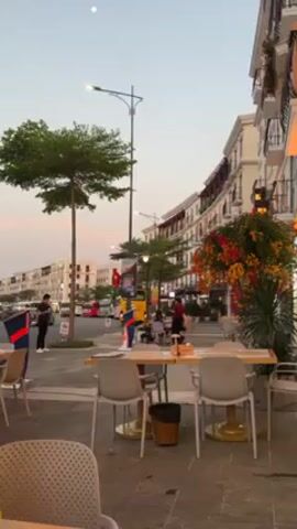 The video presents a static shot of an outdoor café area, likely in a tourist-oriented location. The café tables and chairs are arranged on a paved plaza or wide sidewalk. Several tables are empty, while others seem to be prepared for service with table settings and promotional materials.

Behind the café area is a street with light traffic, including motorcycles and buses. A few people are walking and standing on the sidewalk.

The backdrop consists of multi-story buildings with architectural features, such as balconies and decorative details. The buildings appear to be newly constructed or well-maintained. The sky is lightly colored, suggesting it is either dawn or dusk. The lighting conditions are soft.

The video primarily captures a serene urban scene focused on the café setting and the surrounding environment.
