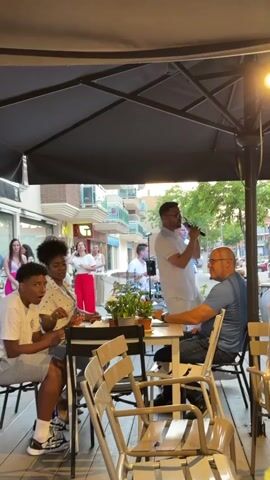 The scene captures an outdoor dining area where a band is performing for the patrons. Two male singers in white shirts stand with microphones. A male musician to their left is playing a metal wind instrument and standing in front of a drum set. Several customers are seated at tables under a large black patio umbrella. One table is occupied by a woman in a polka dot top, a boy in a white shirt and shorts, and a bald man in a blue shirt. The boy appears to be eating something, while the man is looking at the band and resting his chin on his hand. Other patrons are seated at tables in the background. The setting seems to be a street-side cafe, with buildings visible across the street. The atmosphere is lively, with music and people enjoying themselves.