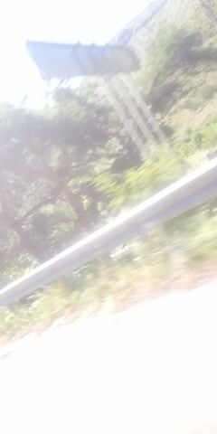 The video shows a view from inside a vehicle, looking out the window. The landscape consists of green hills, trees, and a road with a guardrail. The sun is shining, and the sky is blue. The camera is angled vertically, giving the impression of looking down on the scene. There is some reflection on the window, making it difficult to see certain details clearly.