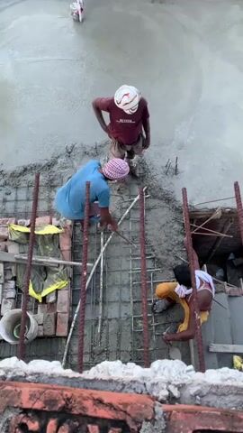 An overhead shot shows three construction workers on a concrete surface. One worker stands near the top center of the frame, wearing a maroon t-shirt, plaid shorts, and a white head covering. Another worker in a blue t-shirt and plaid head covering is positioned lower and to the left, bending over and using a tool to work on a metal grid embedded in wet concrete. A third worker, wearing a yellow garment and a white head covering, kneels to the right, also working with the metal grid and concrete. Reinforcement bars and construction materials are visible around the workers. The scene appears to be a rooftop or elevated construction area.