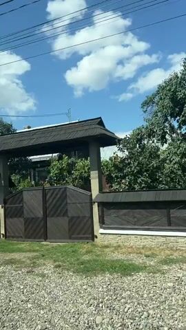The video shows a view from a moving vehicle, likely a car, traveling along a road. The camera is positioned to capture the passing scenery. In the foreground, there is gravel and grass. A dark wooden fence with stone pillars separates the road from properties behind it. Behind the fence, several houses are visible, each with different architectural styles and colors, ranging from white to yellow and beige. Some houses have balconies, and trees and foliage are interspersed between them. The sky is blue with scattered white clouds. Power lines are visible against the sky. A silver car is parked in front of one of the houses. The video captures the exterior of residential properties in what appears to be a rural or suburban setting.
