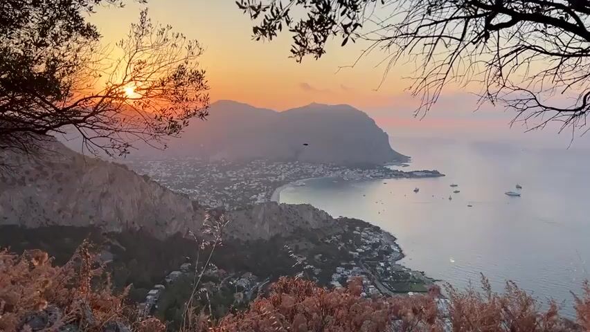A high-angle, wide shot captures a scenic coastal town nestled between rugged mountains and the calm sea during sunset. The sun is partially obscured by a mountain peak on the left, casting a warm orange and yellow glow across the sky and illuminating the town with soft light. The town, characterized by its dense cluster of buildings, stretches along the coastline, with a bay in the center where several boats are anchored. Lush green trees and shrubs dot the landscape, particularly on the slopes of the mountains. In the foreground, dried grasses and tree branches frame the view, adding depth and texture to the composition. The overall atmosphere is serene and picturesque, highlighting the natural beauty of the location at dusk.