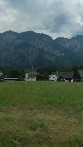 A landscape view of a grassy field with some houses and mountains in the background. A sign with German text and logos is visible on the left side of the frame. The sky is cloudy. The video appears to be taken from a moving vehicle.