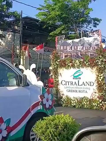 The video shows a parade float decorated with flowers and images of houses. A large sign on the float reads "CITRALAND GRESIK KOTA". There are people on the float, including two individuals sitting and wearing traditional attire, and a large colorful statue of a person at the front. In the background, there are buildings, trees, power lines, and a street with some people on motorcycles. The Indonesian flag is also visible, draped over a structure. The scene appears to be a daytime event, likely a celebration or promotion.