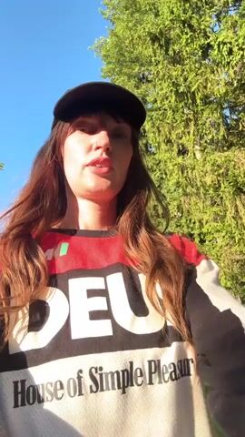 This is a first-person video taken by a woman wearing a black baseball cap with "Deus Tokyo Japan" and a black, red, and white long-sleeved shirt with "Deus House of Simple Pleasure" printed on it. She is walking on a dirt path outdoors, with trees and foliage in the background. To her right and slightly behind her is another woman wearing sunglasses and a dark sweatshirt with "Hard Rock Cafe" on it and green pants. The first woman is speaking to the camera, and the second woman appears to be looking off-camera. The lighting suggests it is daytime, with sunlight filtering through the trees.