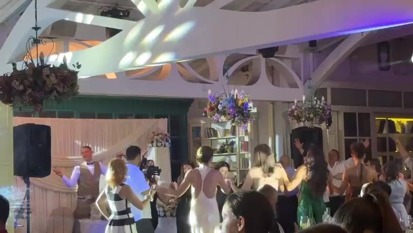 This video captures a lively celebration, likely a wedding or a festive gathering, taking place indoors at night. A group of people, predominantly young adults, are engaged in dancing, forming a circle and holding hands. The atmosphere appears joyous and energetic, with several individuals smiling and actively participating in the dance.

Prominently featured is a man on the left side of the frame, dressed in a vest and bow tie, holding a Bulgarian flag. He seems to be leading or encouraging the dancers. Another man in the circle is wearing a white shirt and appears to be a central figure in the dance. The female dancers are dressed in various attire, including a white backless dress and a green dress.

The setting is a well-decorated indoor venue. Overhead, ornate chandeliers with floral arrangements hang from the ceiling, adding a touch of elegance. The walls are adorned with white drapes and some subtle lighting effects, including green and purple hues, enhance the festive mood. In the background, shelves filled with books are visible, suggesting a potentially eclectic or historic venue. The presence of speakers indicates that music is a key element of the event.

The time of day is clearly nighttime, as evidenced by the indoor lighting and the absence of natural daylight. While specific weather conditions are not discernible, the indoor setting suggests protection from any external elements. The location context of Krepost, Bulgaria, provides a geographical reference for the event. The overall mood is one of celebration and camaraderie. No visible text is present in the video.